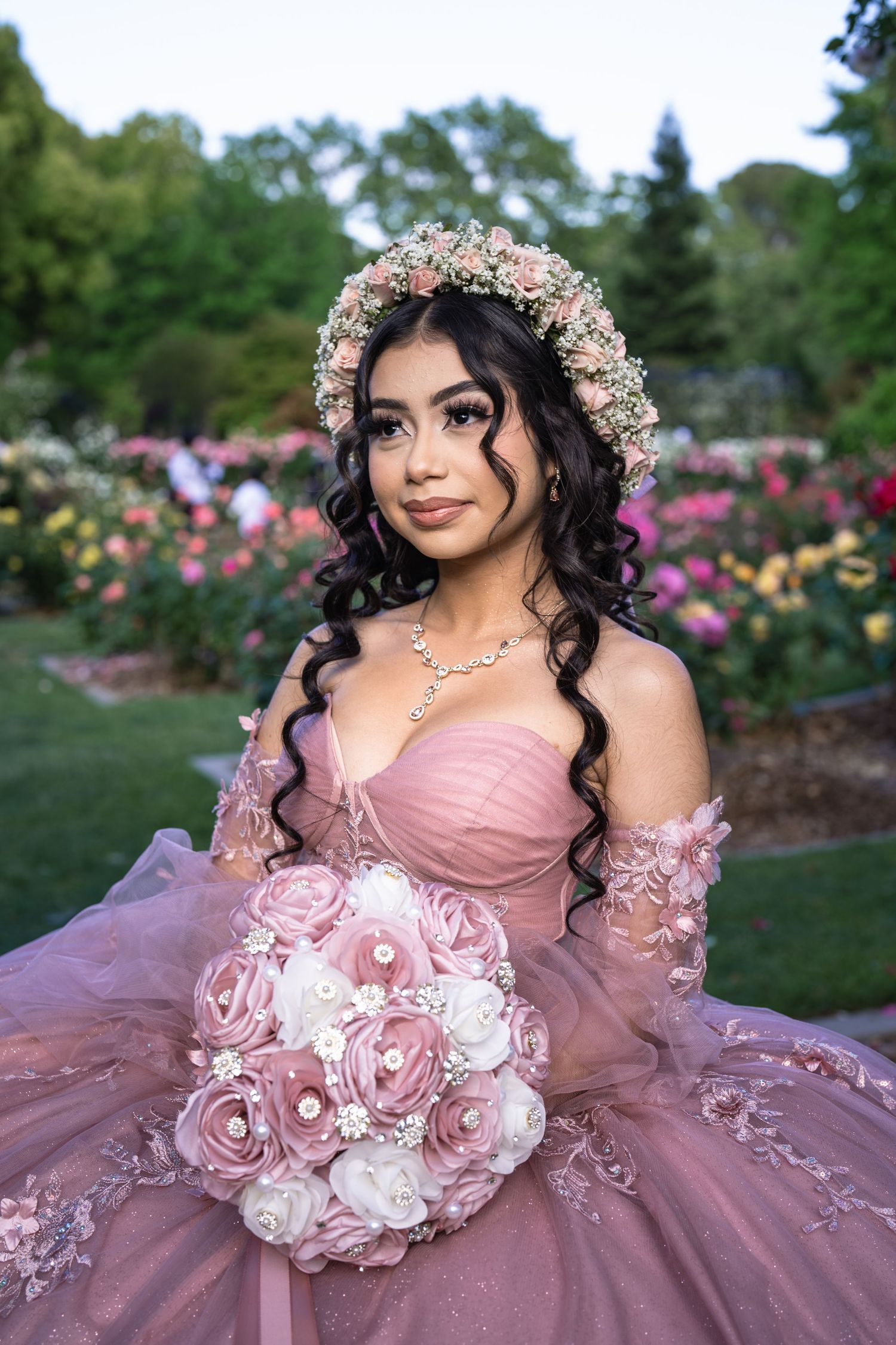 Young woman in pink quinceañera gown with floral crown holds bouquet in blooming garden.
