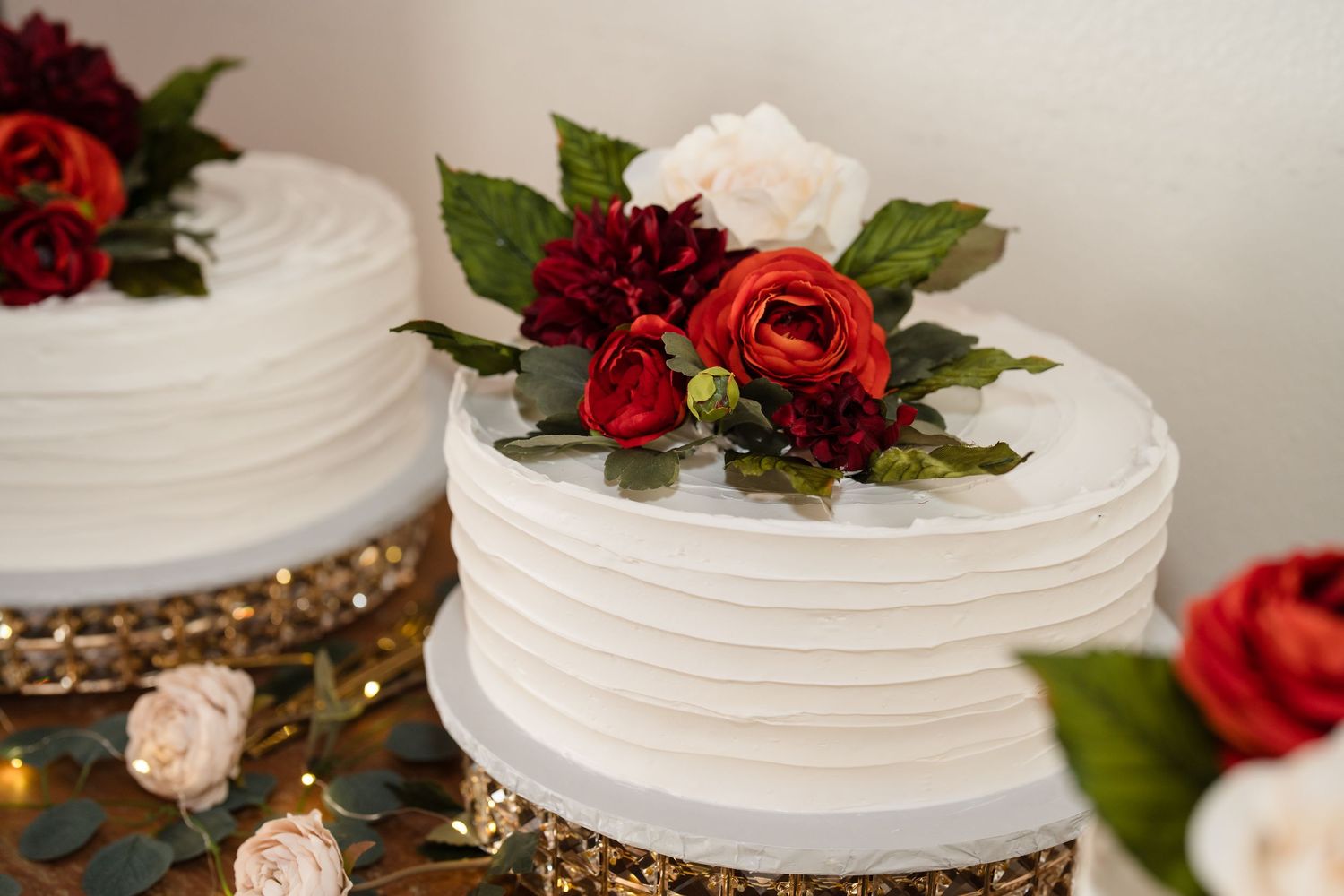 White wedding cake with red and white floral topper on gold stand.