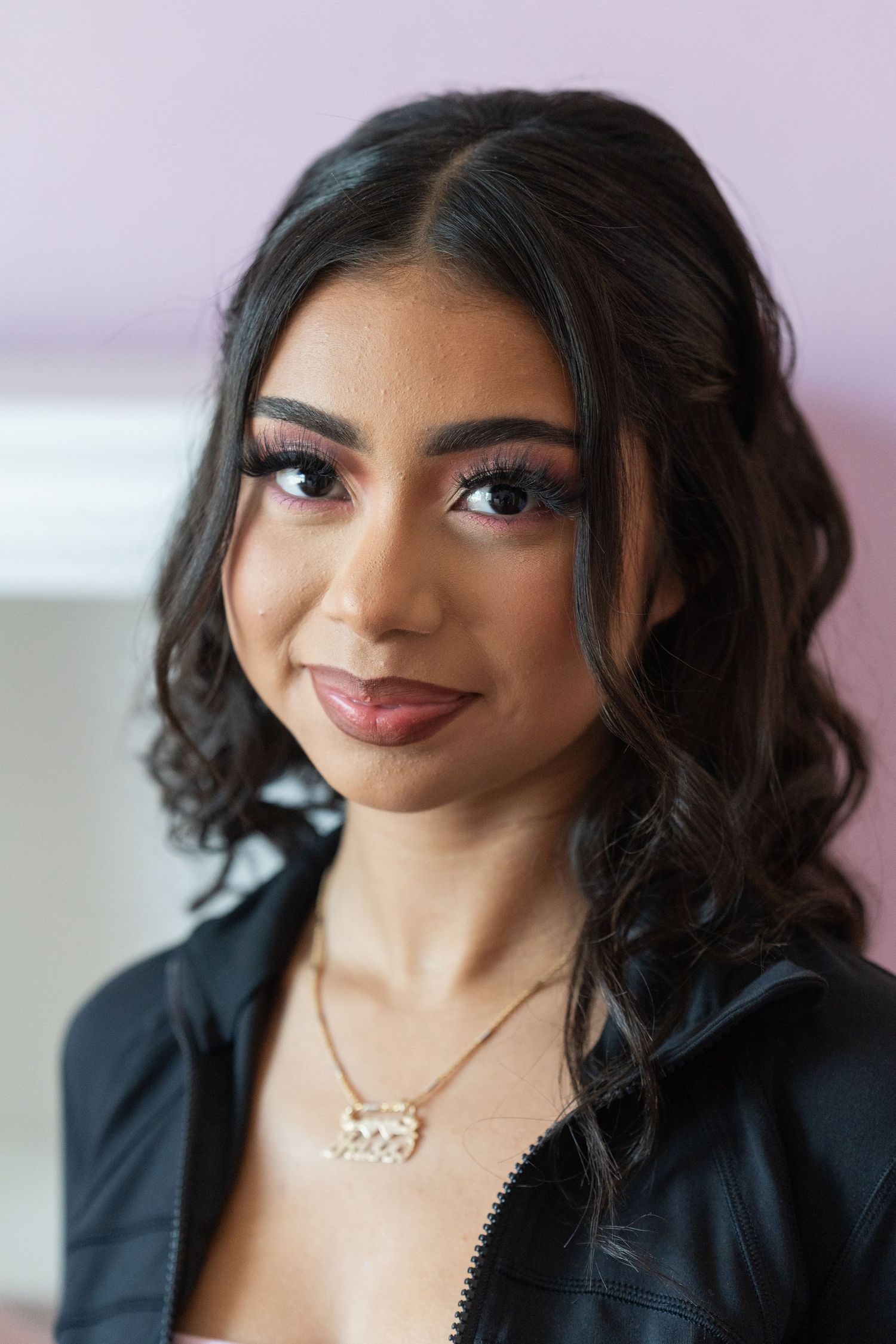 Young woman with glamorous makeup and dark curly hair wearing a black jacket and gold nameplate necklace.