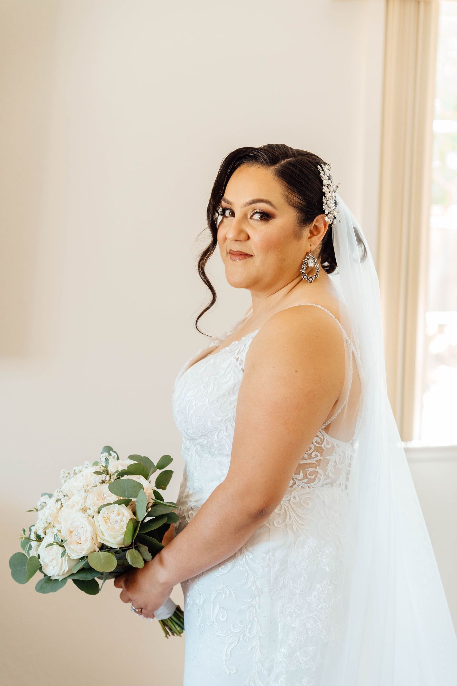 Beautiful bride in lace wedding gown holding white rose bouquet with veil and crystal hair accessories.