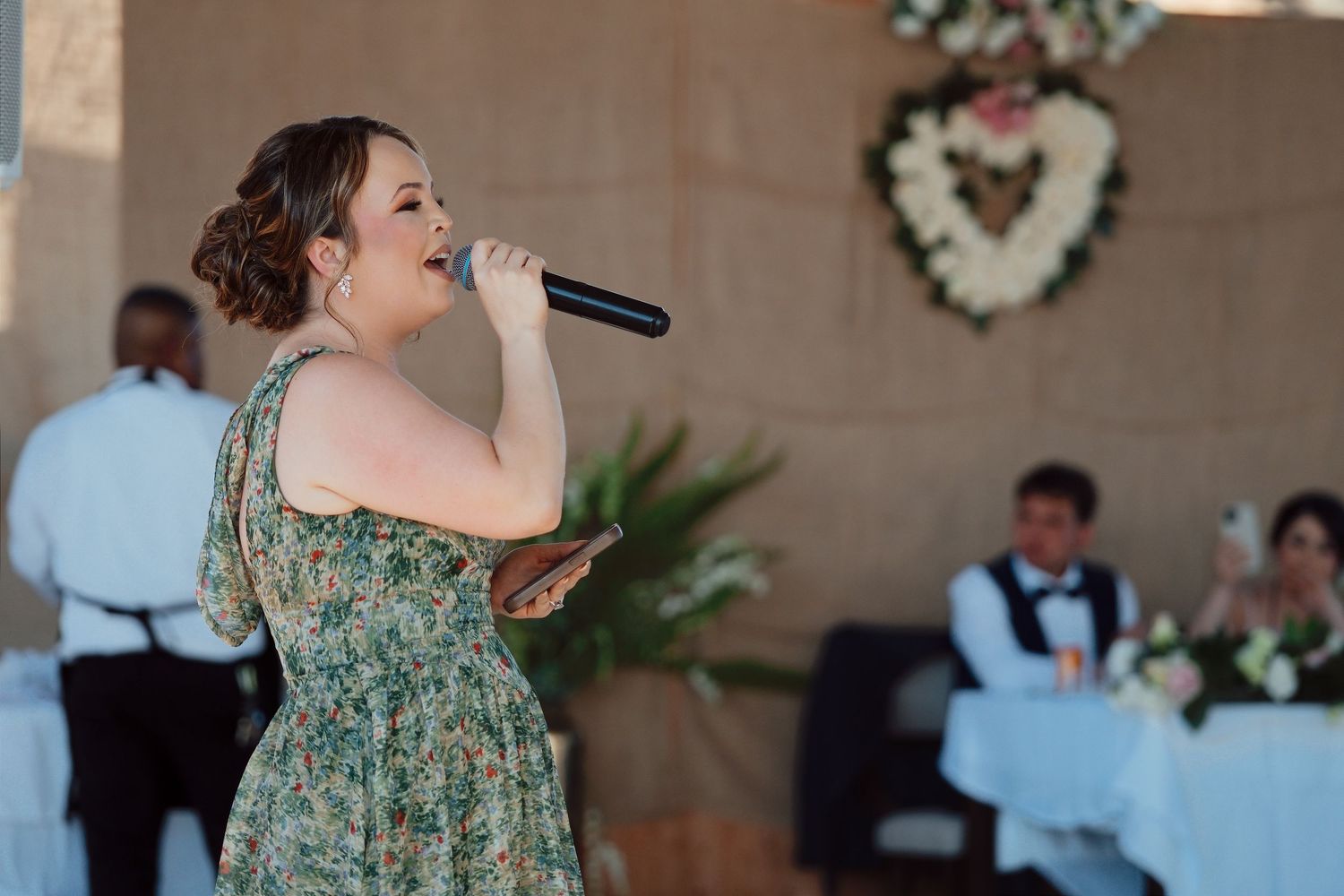 Woman in floral dress singing into microphone at outdoor wedding reception with guests seated in background.