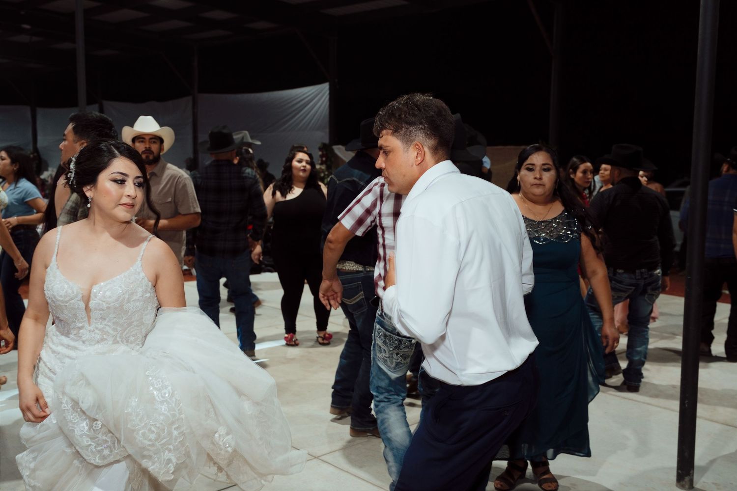 Bride in white wedding gown dancing with a man in white shirt at a wedding reception with guests watching.