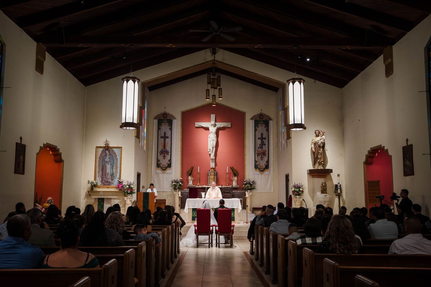 Catholic church interior with congregation seated during Mass, featuring crucifix altar and stained glass windows.