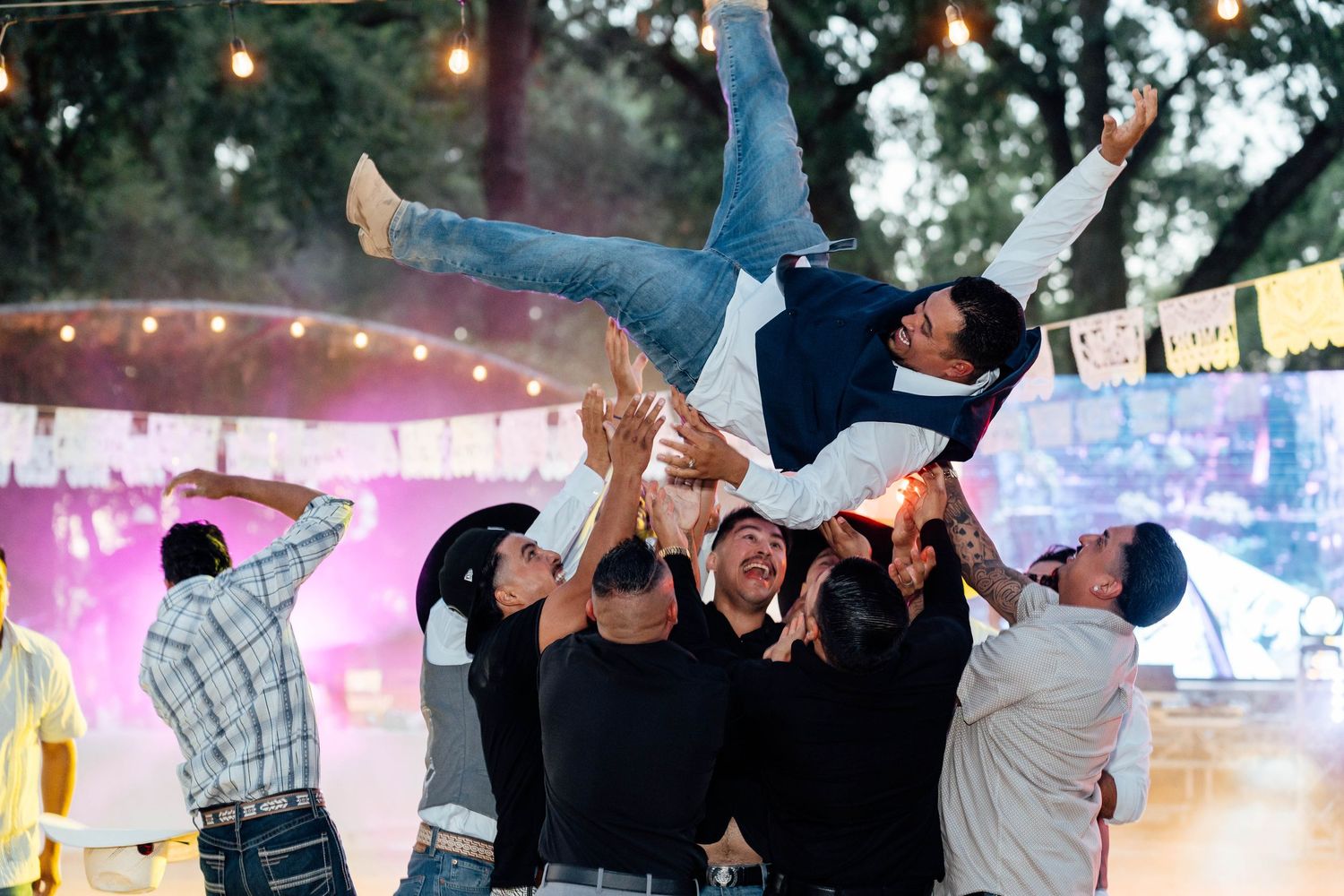 Groomsmen tossing groom into the air at outdoor wedding celebration under string lights at night.