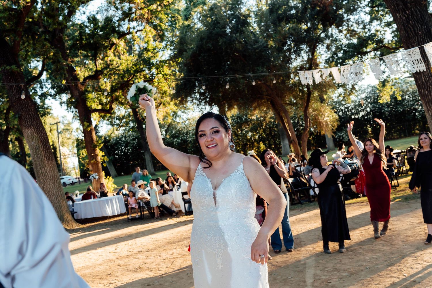 Bride in white dress dancing joyfully outdoors at wedding reception under trees with string lights.