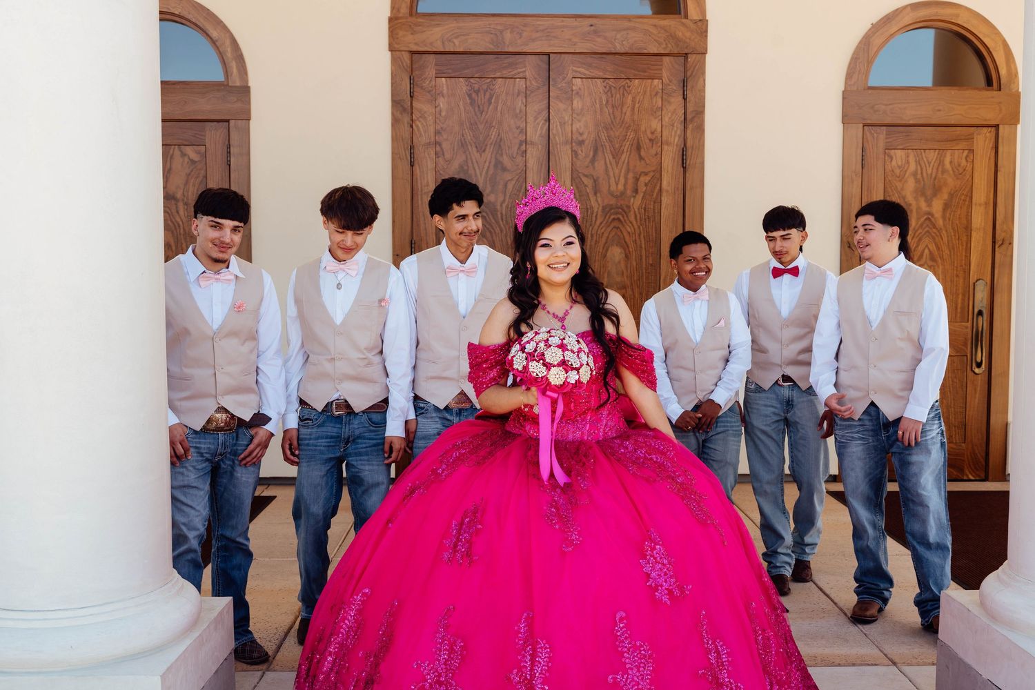 Quinceañera girl in hot pink gown poses with chambelanes in beige vests outside church entrance.