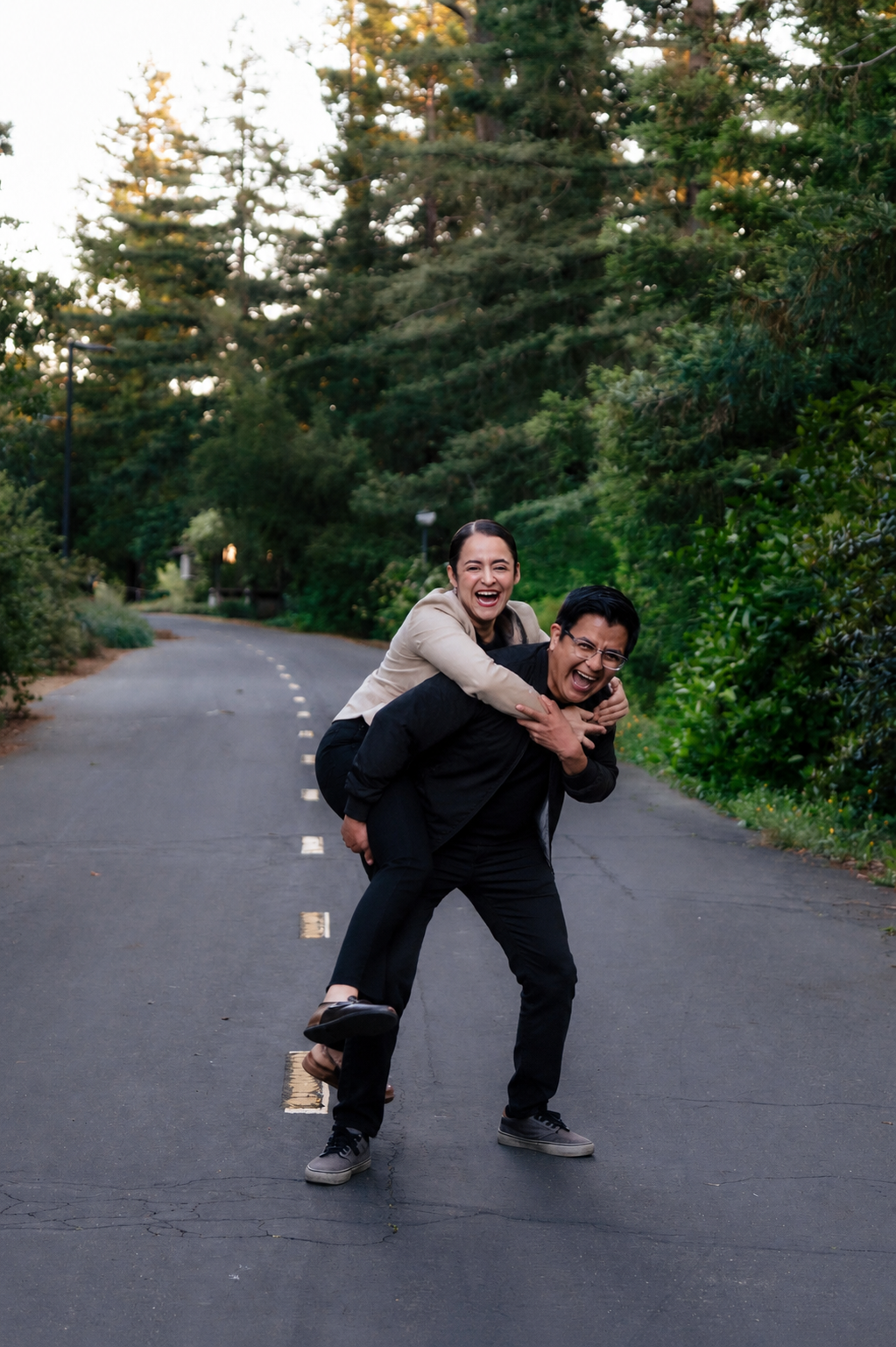 Laughing couple plays piggyback on a tree-lined road, woman on mans back in a joyful outdoor portrait.