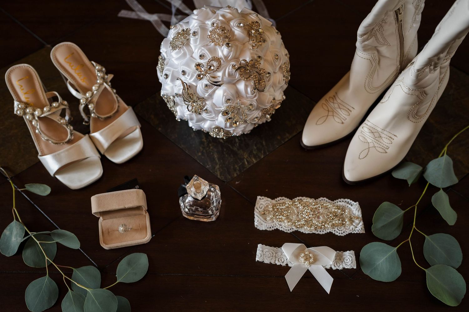 Bridal flat lay featuring white bouquet, gold sandals, cowboy boots, ring box, perfume, and lace garter on dark wood.