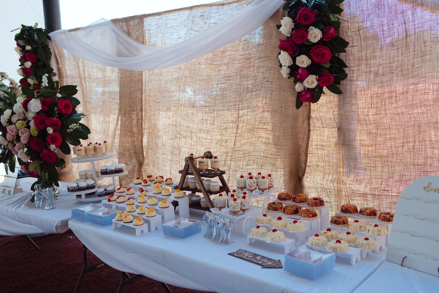 Elegant wedding dessert table with white linens, assorted sweets, and red and white floral arrangements on burlap backdrop.