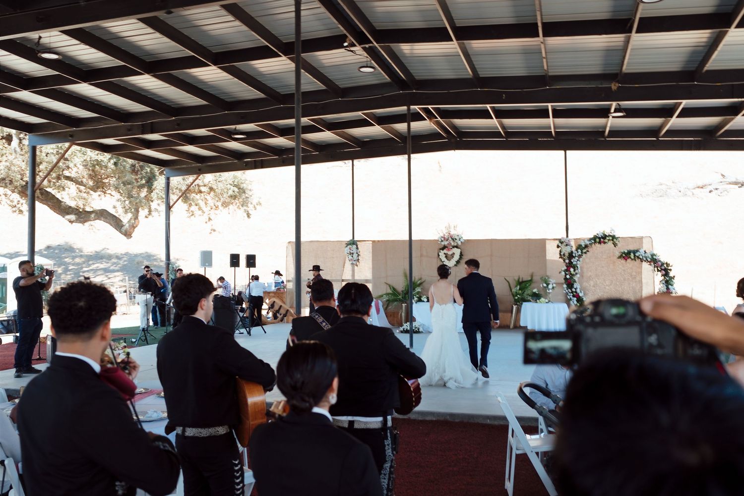 Outdoor wedding ceremony under a covered pavilion with guests seated, couple at the altar, and a cameraman filming the event.