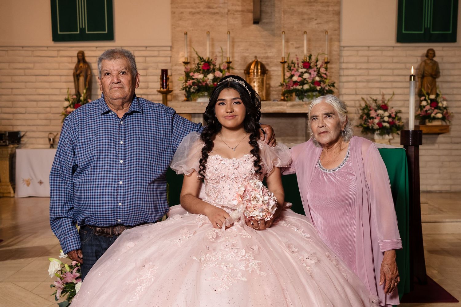 Young girl in pink quinceañera gown poses with elderly couple near church altar adorned with floral arrangements.