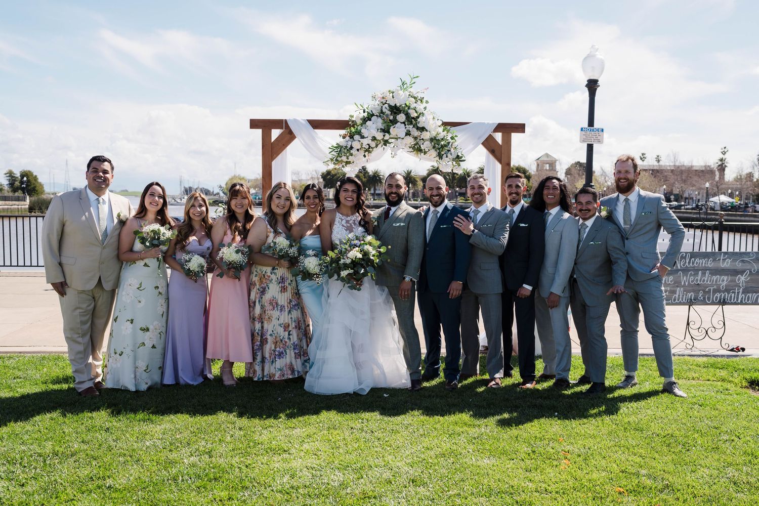 Wedding party poses outdoors near a floral arch, bridesmaids in pink and the couple at center on a sunny day.