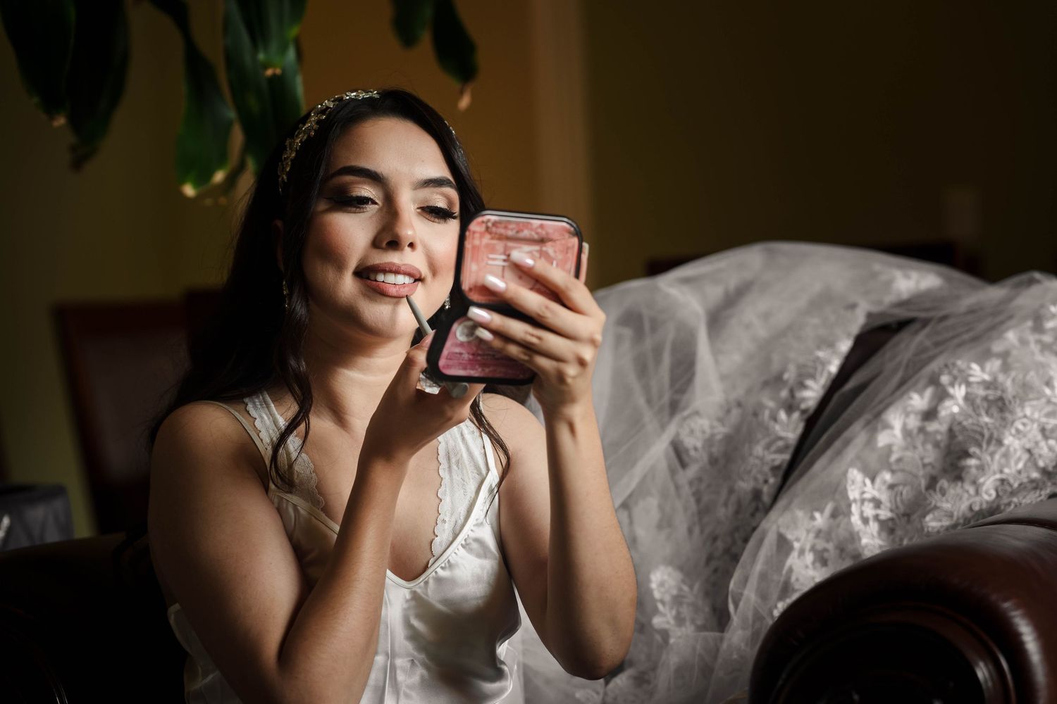 Bride in white wedding gown applying makeup while looking in compact mirror before ceremony.