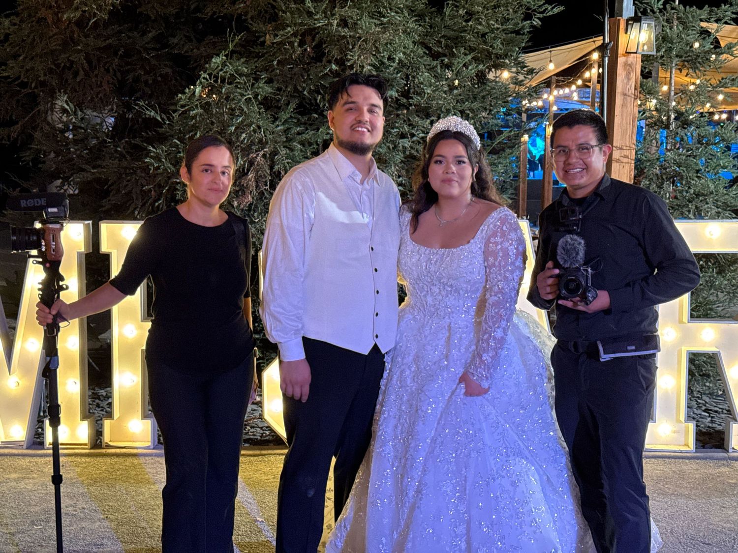 Bride in white gown and groom pose with two guests at an outdoor evening wedding reception.