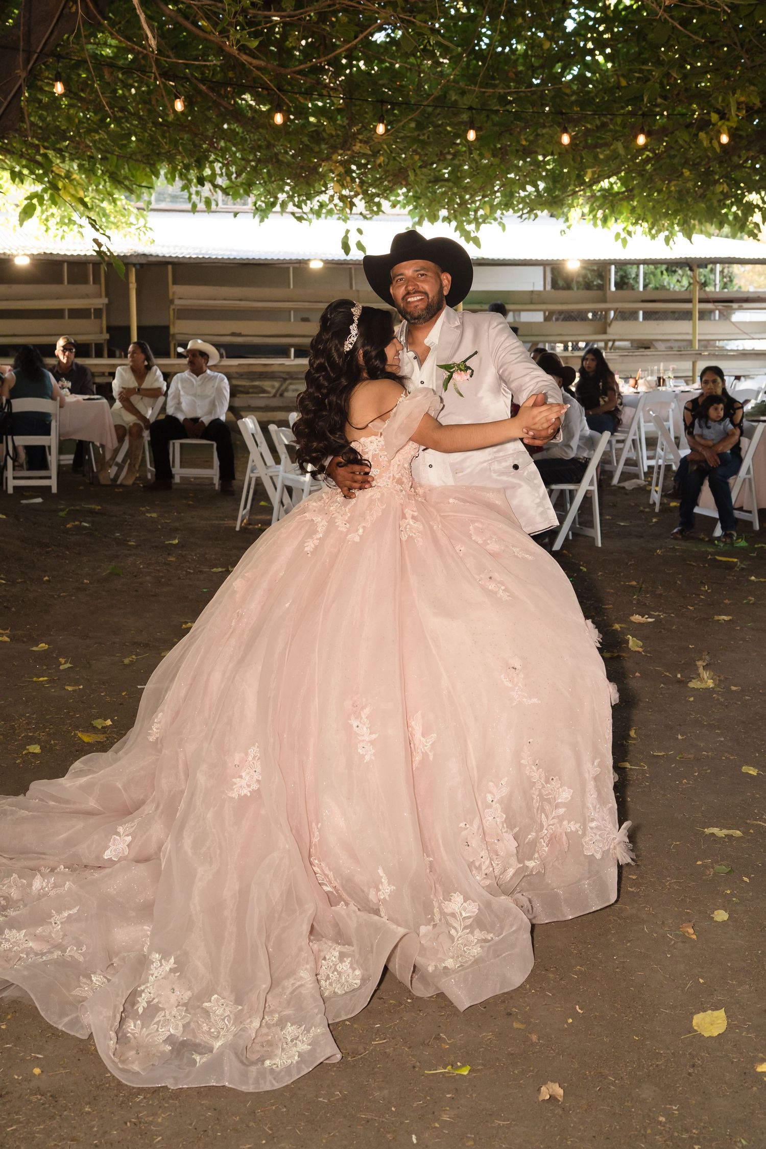 Couple dancing at quinceañera celebration, woman in pink ballgown with floral lace, man in cowboy hat, outdoor venue.