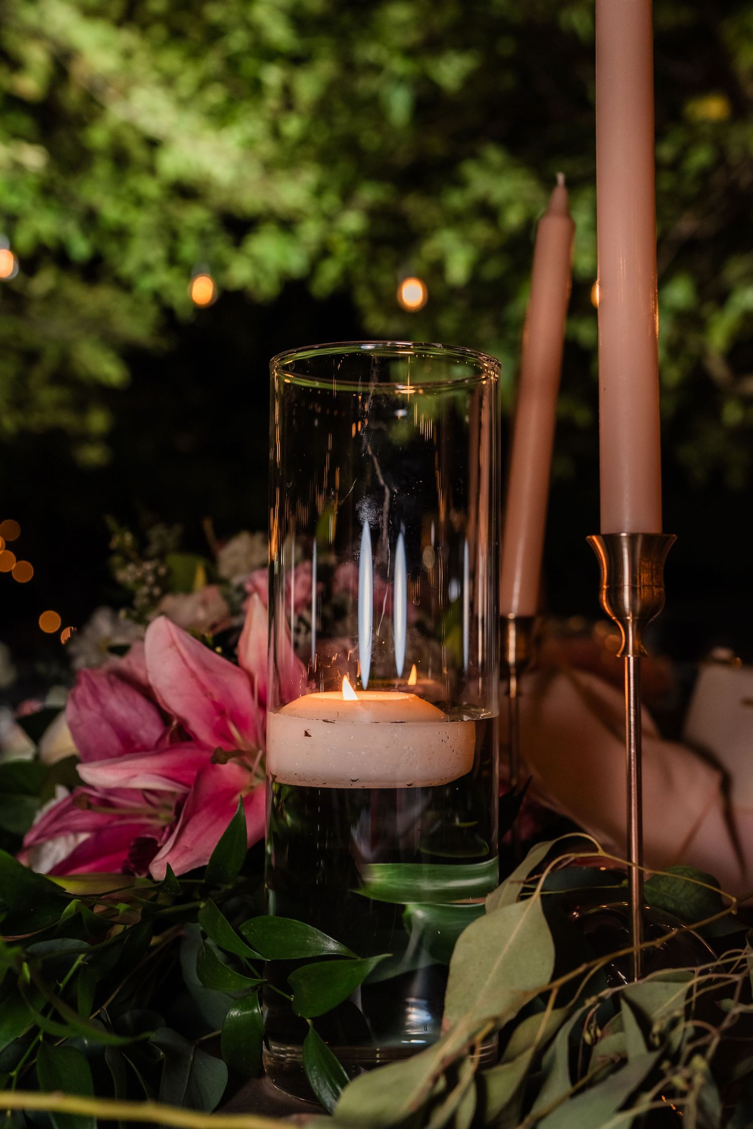 Lit candle in glass hurricane vase surrounded by pink lilies and greenery at night.