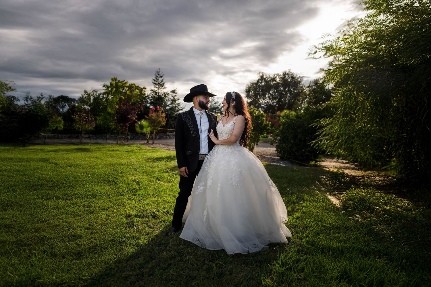 Bride in white ball gown and groom in black tuxedo with cowboy hat share a romantic moment on green lawn at golden hour.