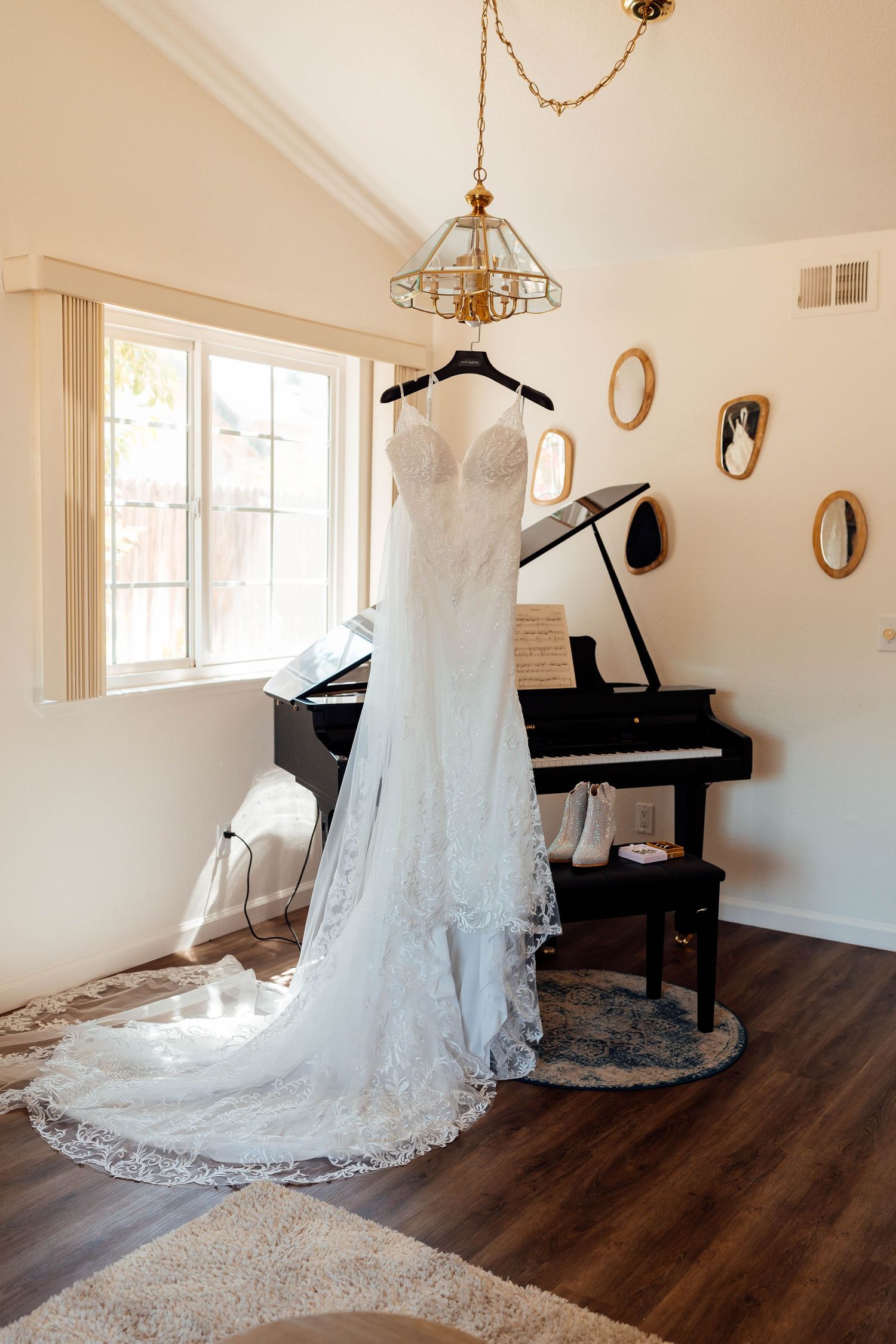 Elegant lace wedding gown with long train hanging from chandelier near black grand piano in bright room.