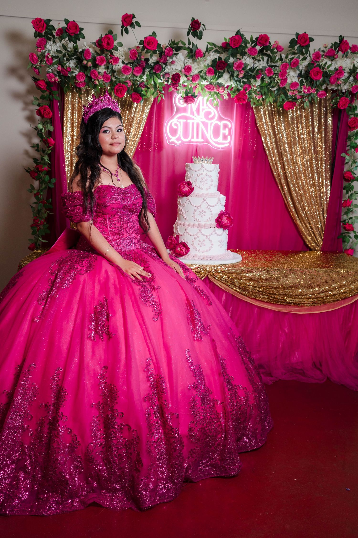 Young woman in a stunning hot pink quinceañera ball gown poses beside a decorated white tiered cake.
