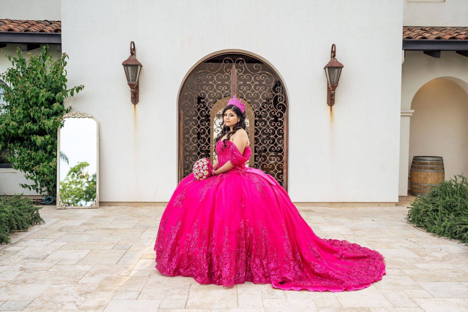 Teen in elaborate hot pink lace quinceañera gown holding bouquet poses at white Spanish-style venue.