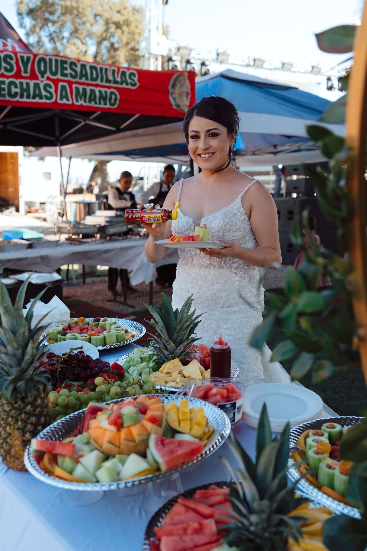 Smiling bride in white gown stands beside colorful fruit display at outdoor wedding reception.
