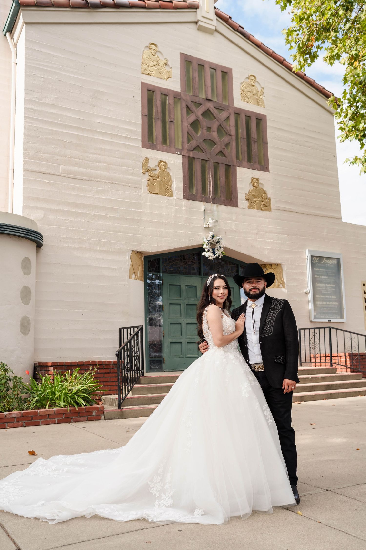 Elegant bride in long white gown with train and groom in black cowboy hat pose before white church facade on wedding day.