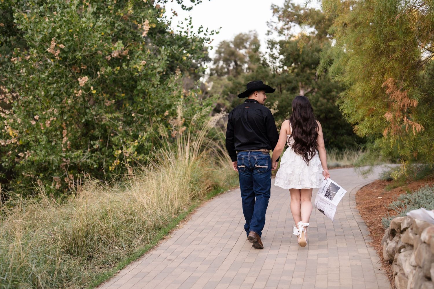 Couple walks hand-in-hand on a garden path, man in cowboy hat and jeans, woman in white dress.