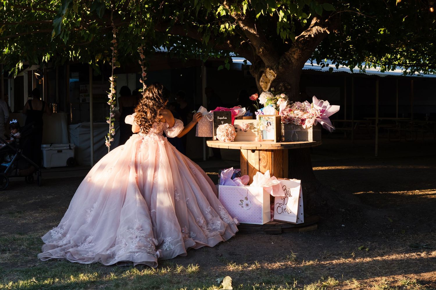 Quinceañera girl in pink ball gown stands at rustic wooden spool gift table under shaded trees.
