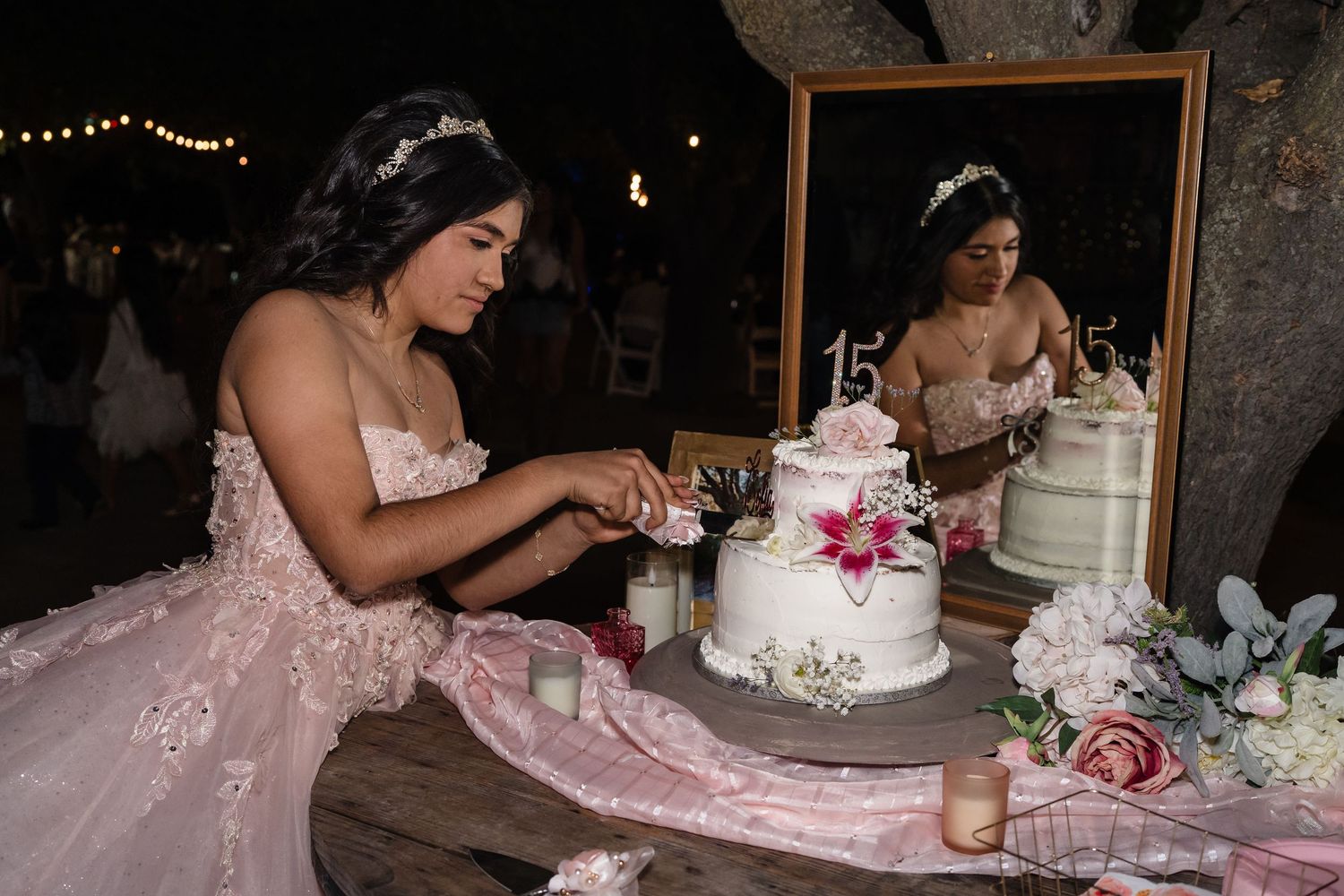 Young woman in pink quinceañera dress cutting her decorated 15th birthday cake at night celebration.