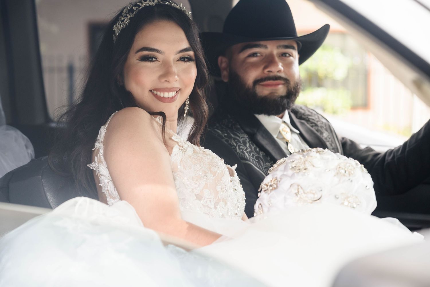 Smiling bride and groom in black cowboy hat sit together in car holding wedding bouquet.