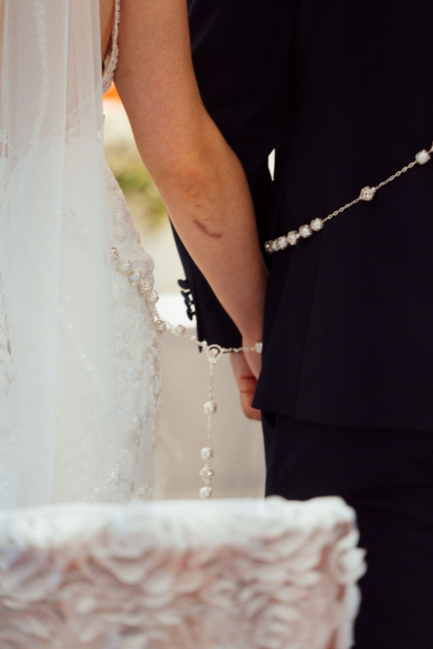 Bride and groom holding hands at wedding altar with lace dress detail and pearl bracelet visible.