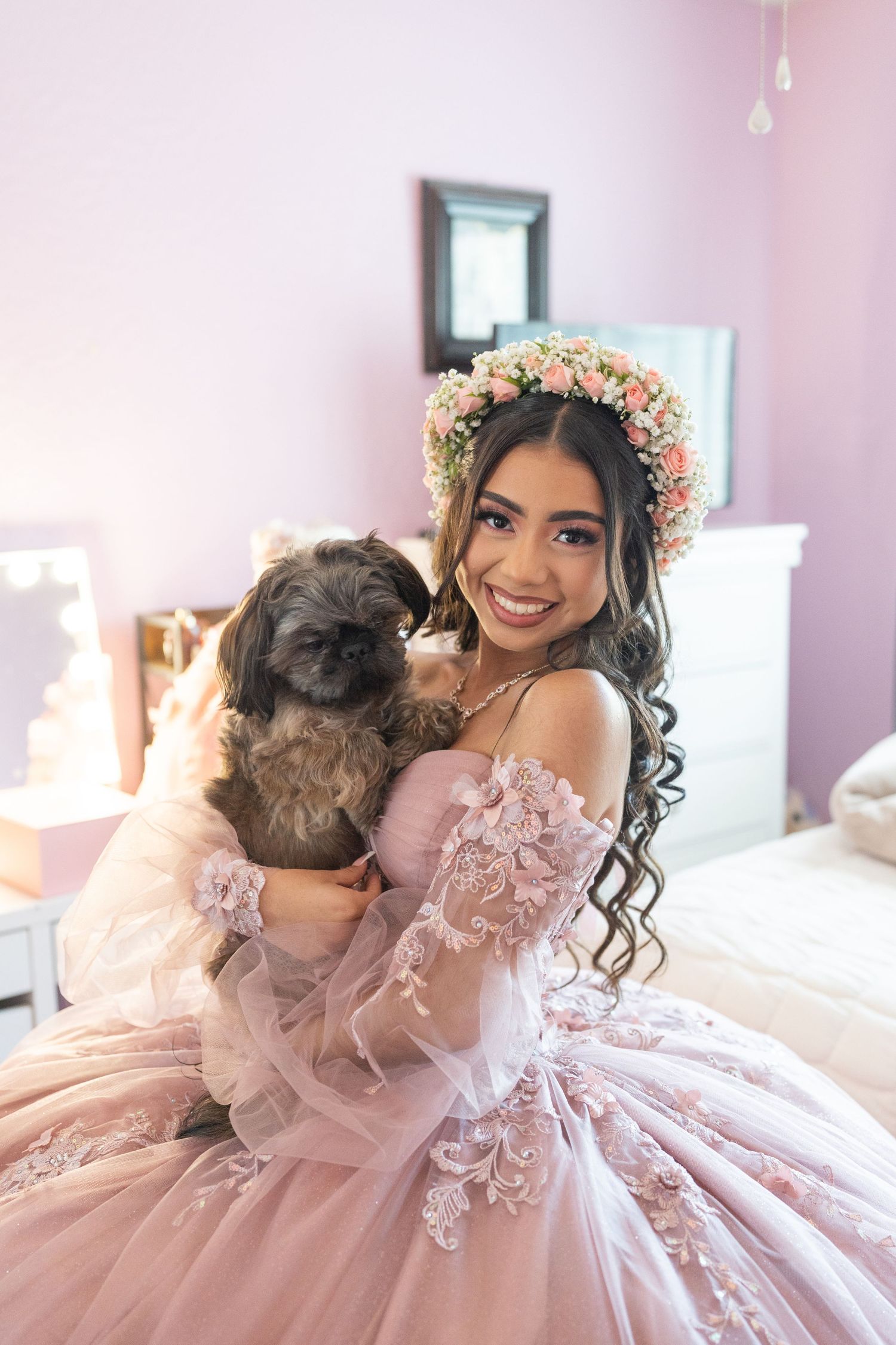 Smiling quinceañera girl in pink ball gown and floral crown happily holding a small fluffy dog.