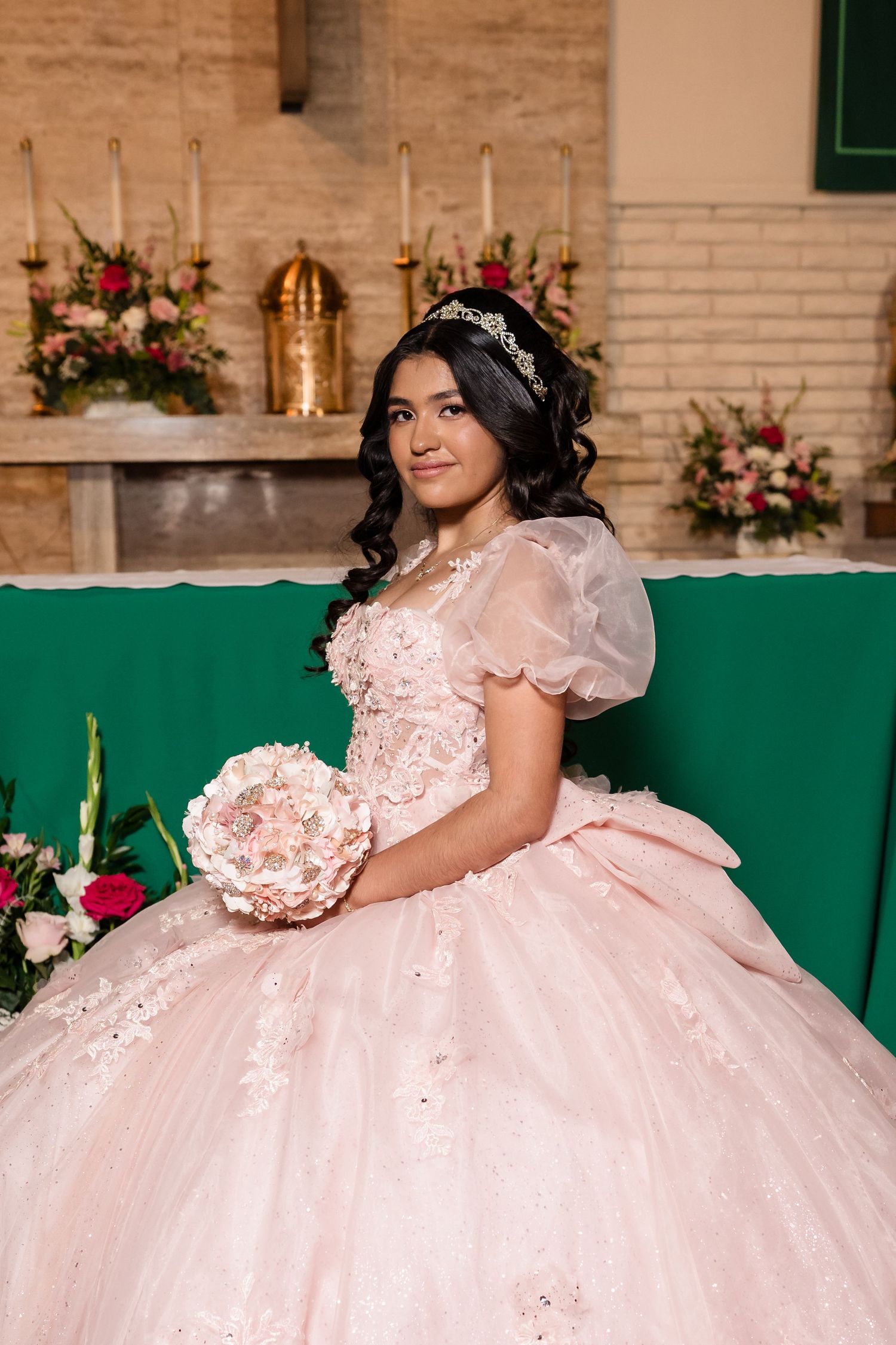 Young woman in pink quinceañera ball gown with tiara holds floral bouquet in decorated church.
