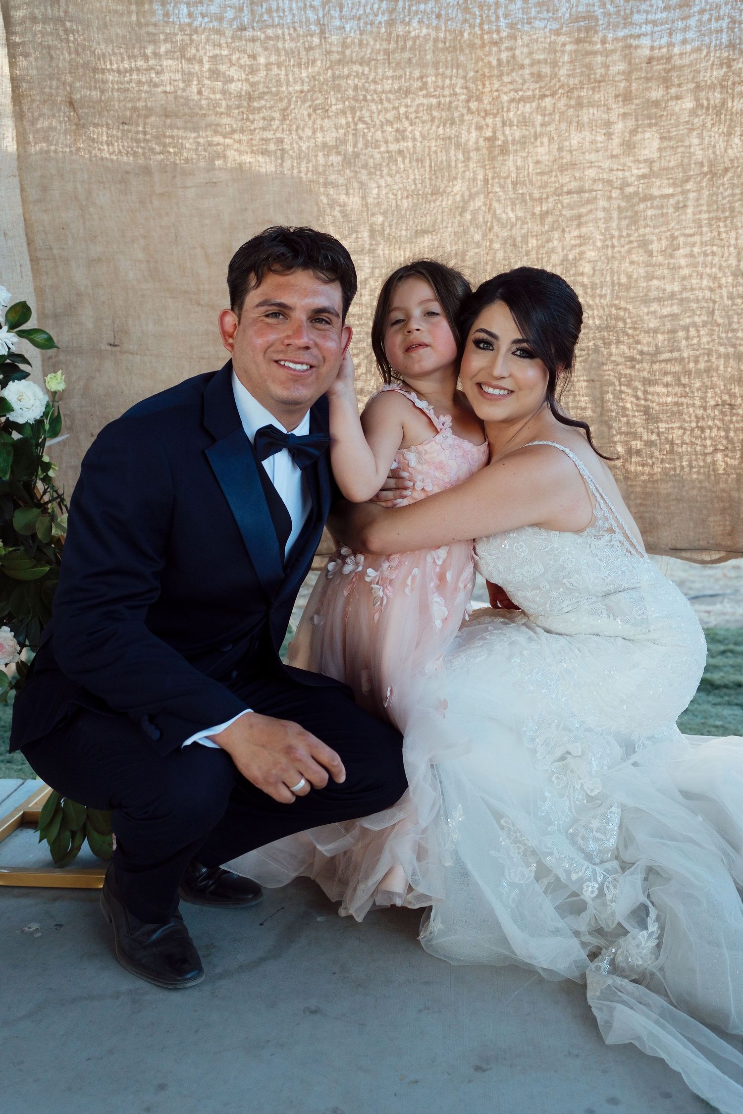 Smiling groom in tuxedo and bride in lace wedding dress pose with young girl in pink dress at wedding.
