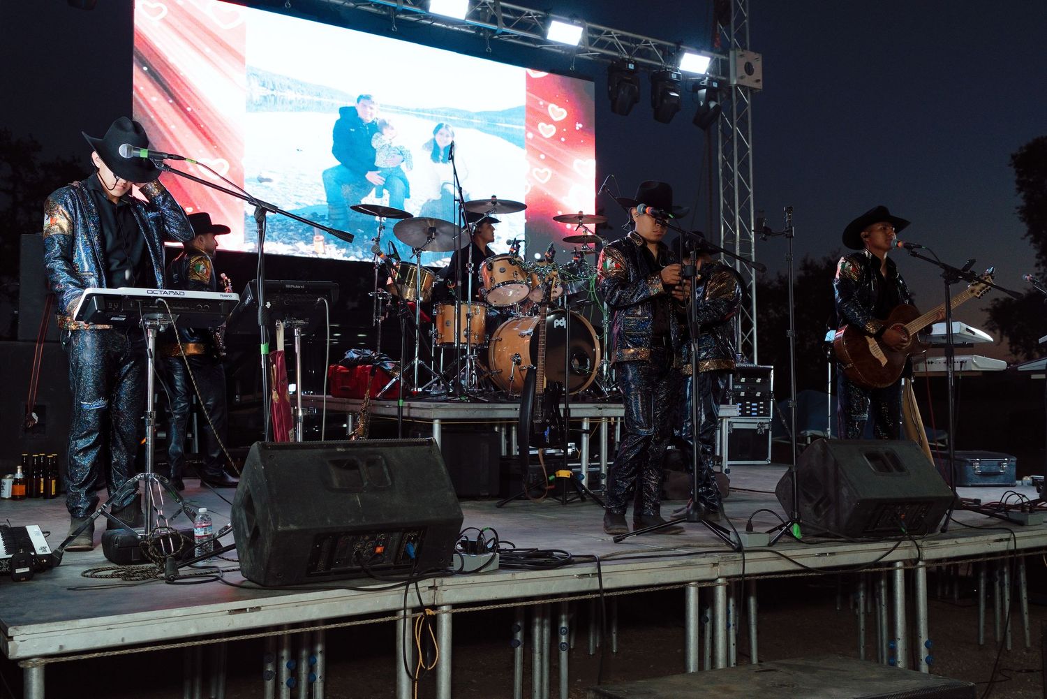 Band performing on outdoor stage at night with large LED screen backdrop and speakers.