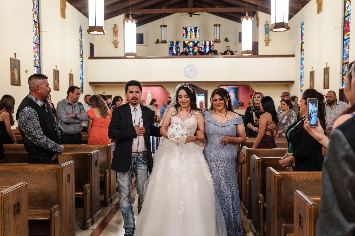 Bride walks down church aisle with two escorts during wedding ceremony, guests watching and photographing the moment.