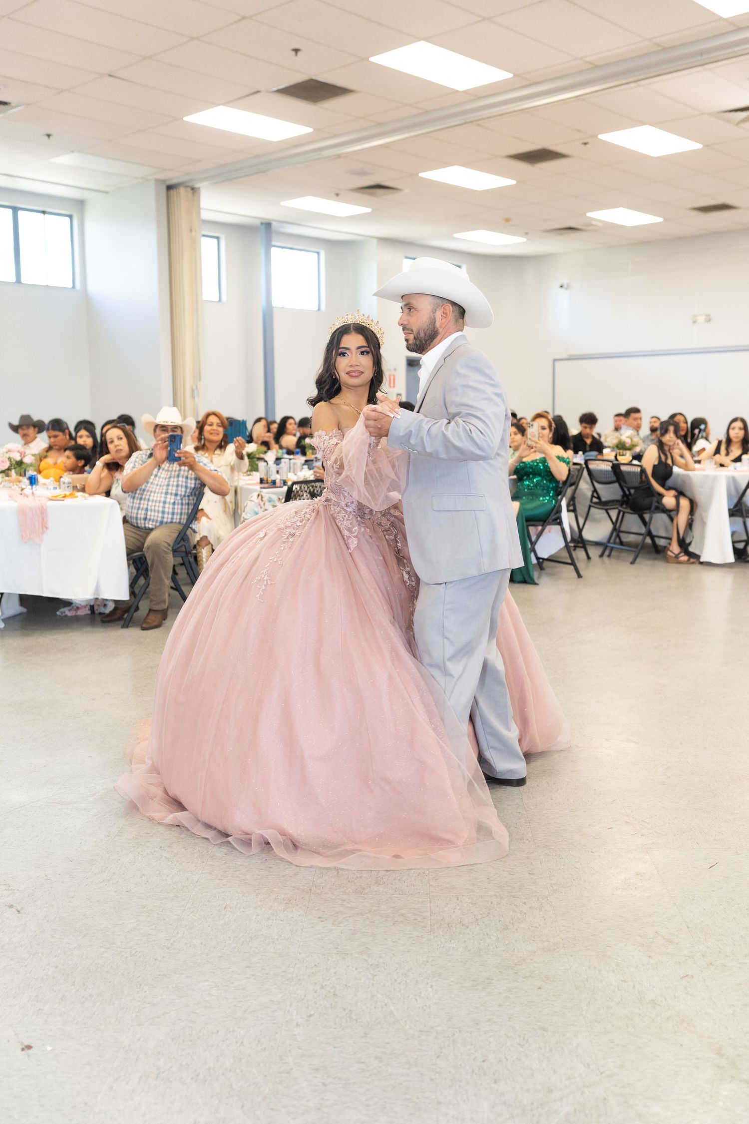A quinceañera girl in a pink ballgown dances with her father in a gray suit at her celebration.