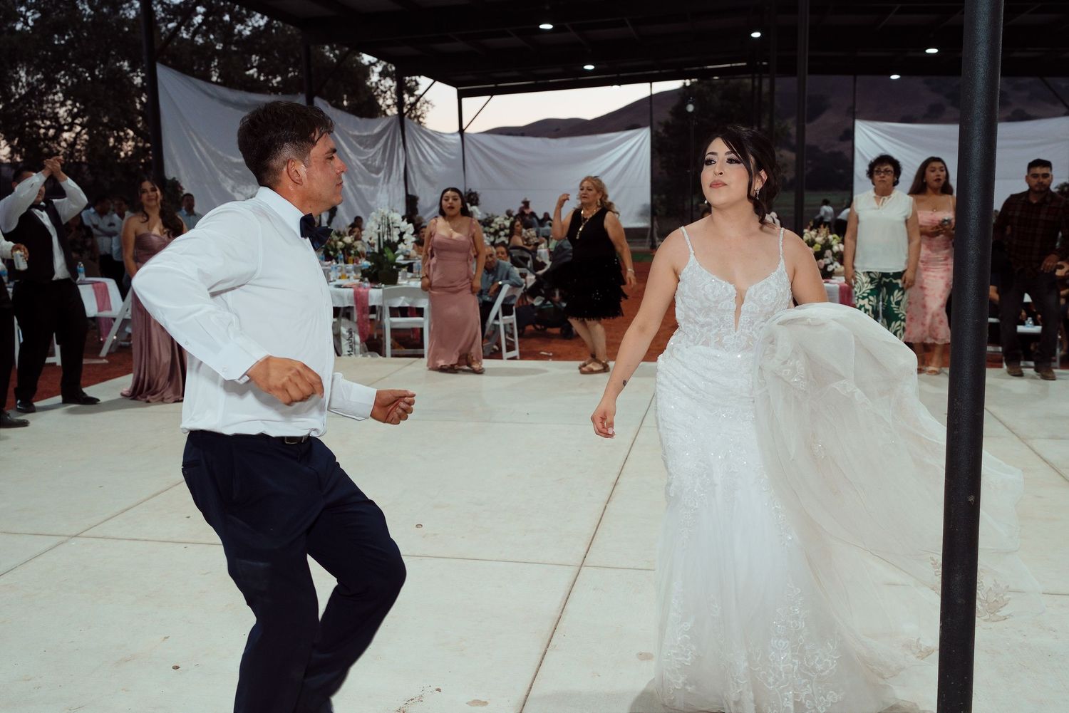 Bride in white gown dances energetically with a guest on outdoor wedding reception dance floor.