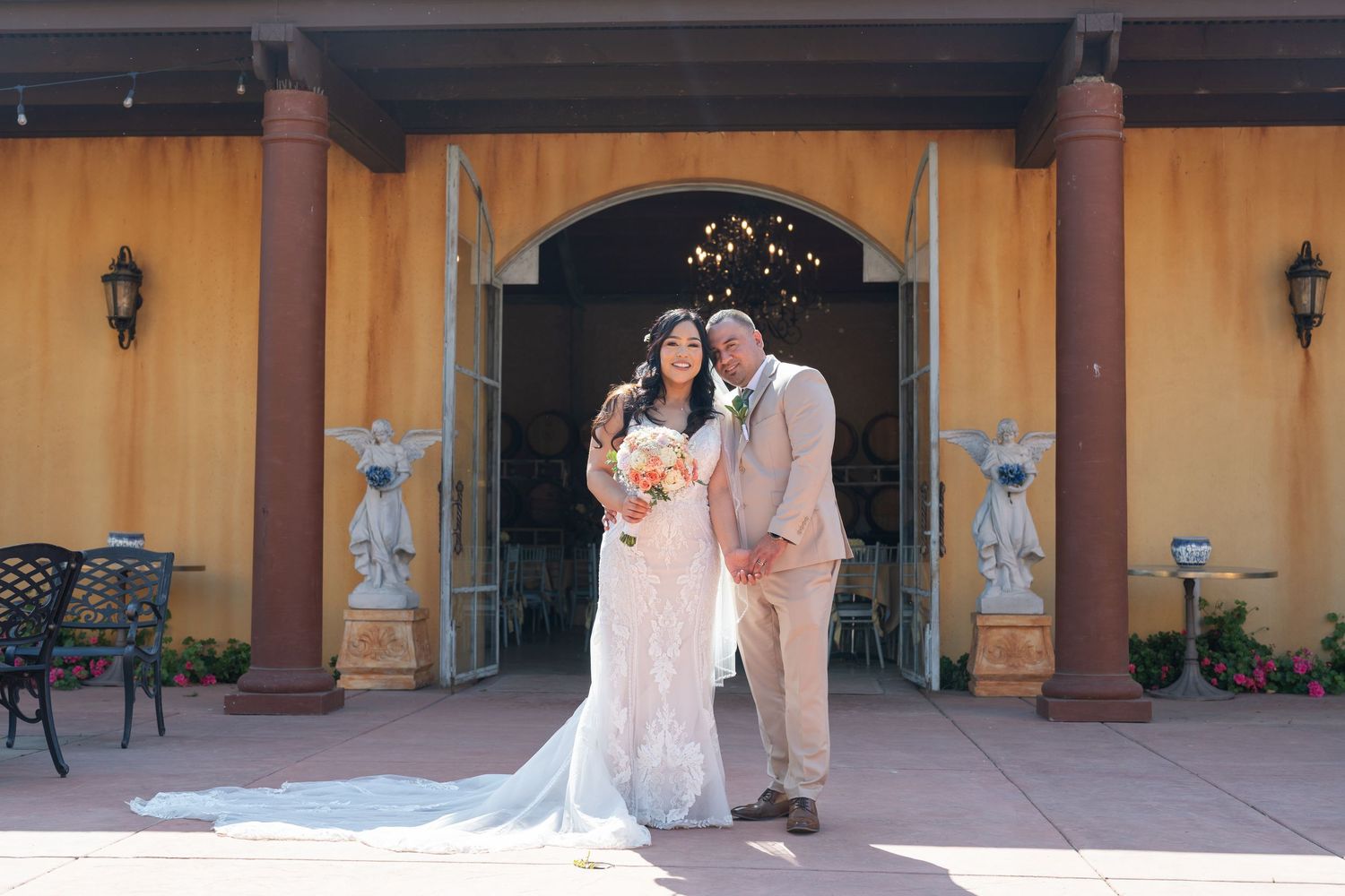 Bride and groom pose together outside elegant yellow venue with columns and string lights on their wedding day.