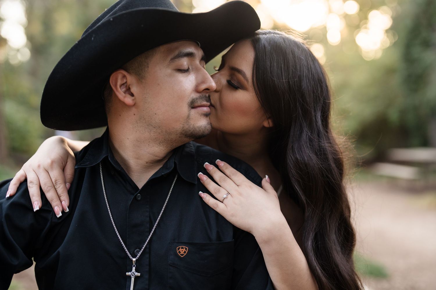 Bride kissing groom on cheek outdoors, groom wearing black cowboy hat and cross necklace, engagement ring visible.