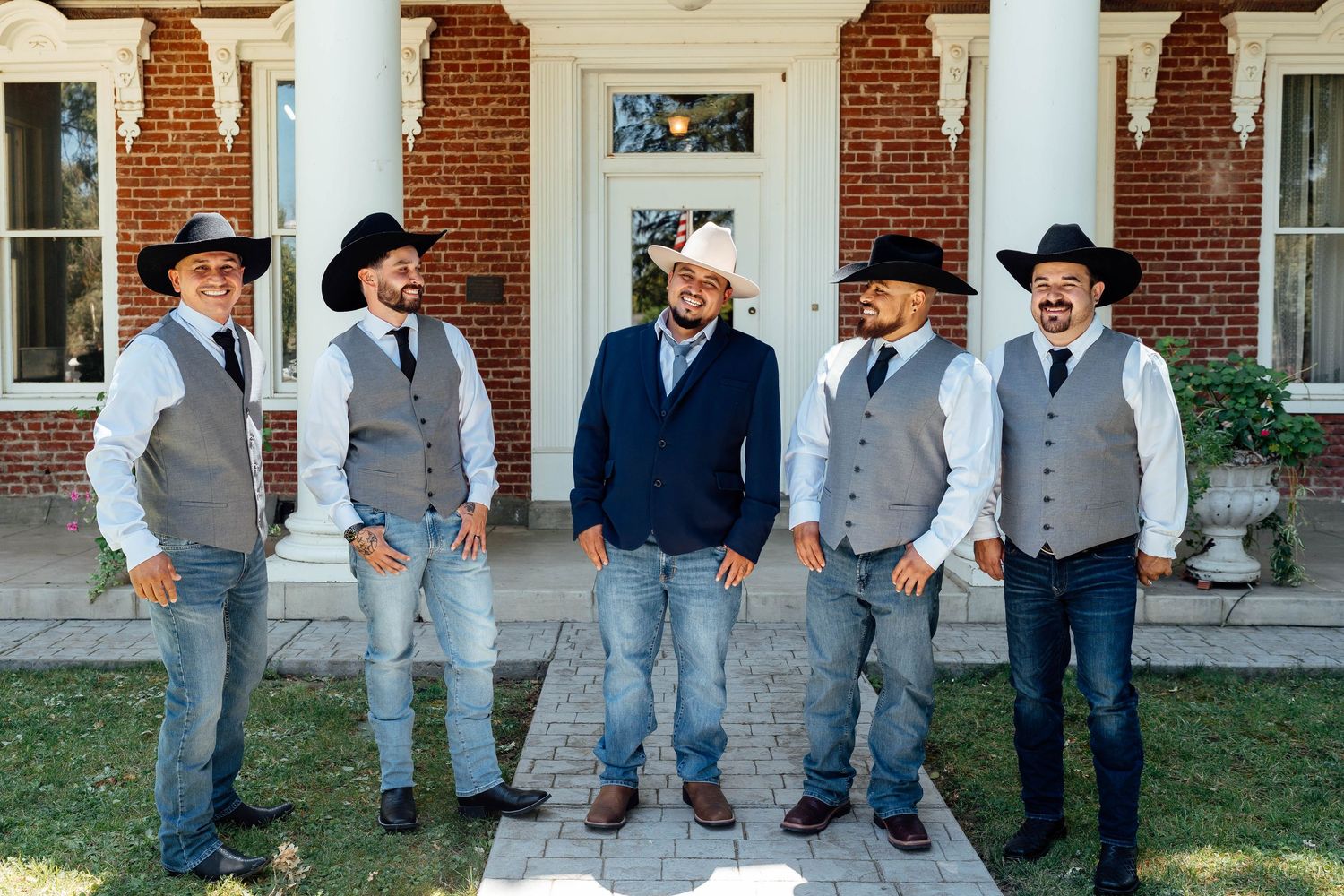 Six groomsmen in cowboy hats, gray vests, and jeans pose outside a brick building.