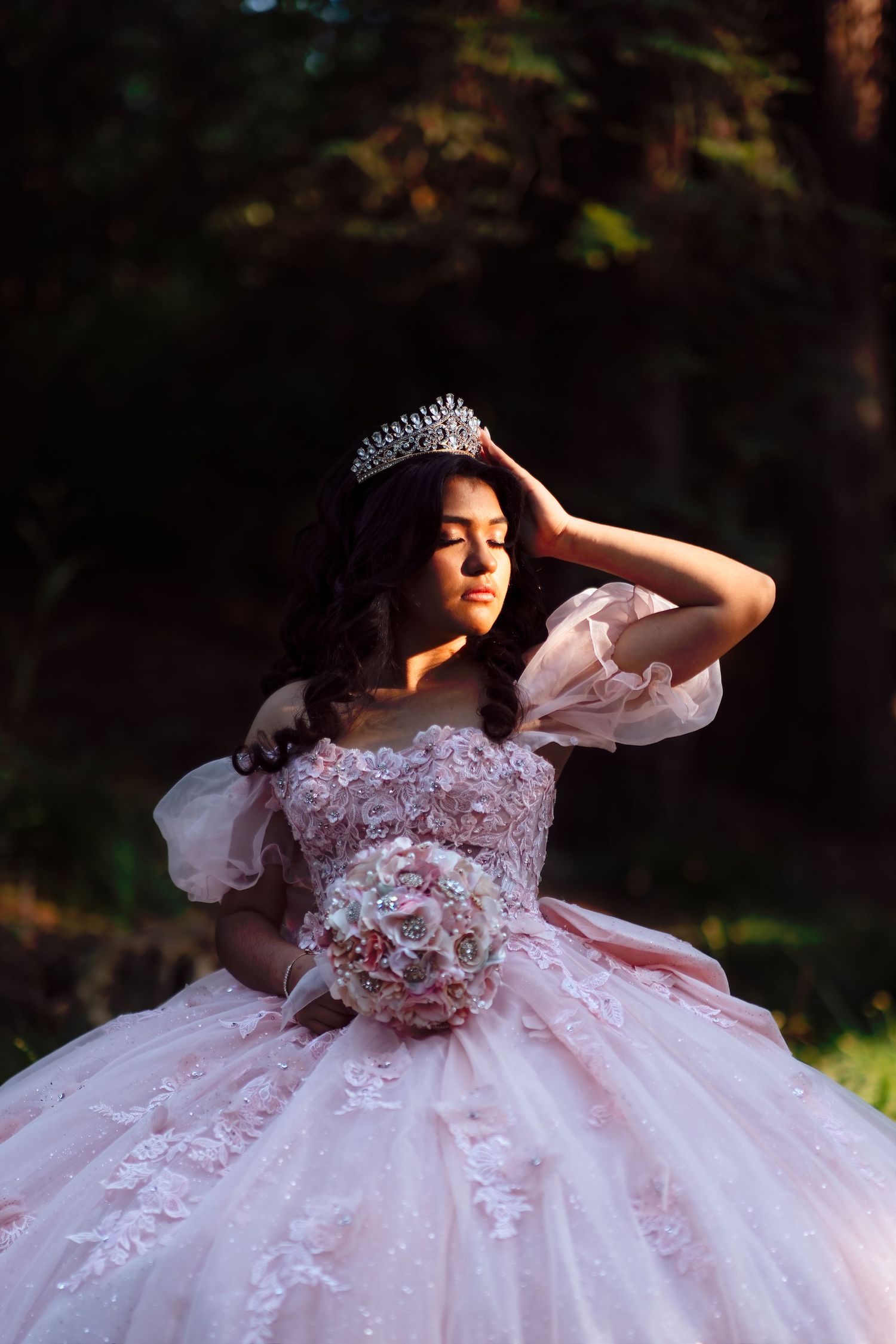 Young woman in pink ball gown with tiara holds floral bouquet outdoors in golden hour light.