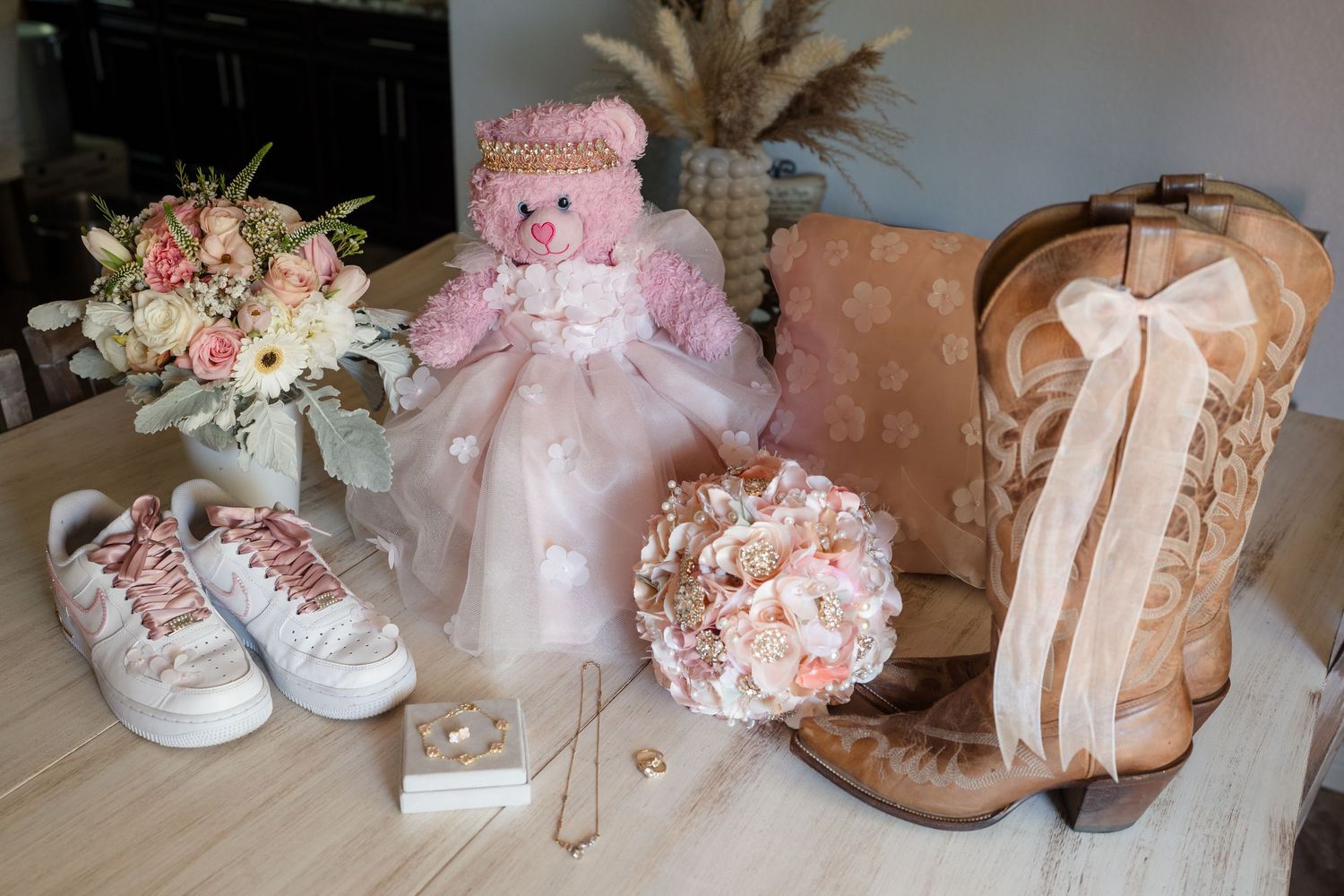 Pink teddy bear, bridal bouquet, cowboy boots, sneakers, and jewelry arranged on a table for a wedding flat lay.