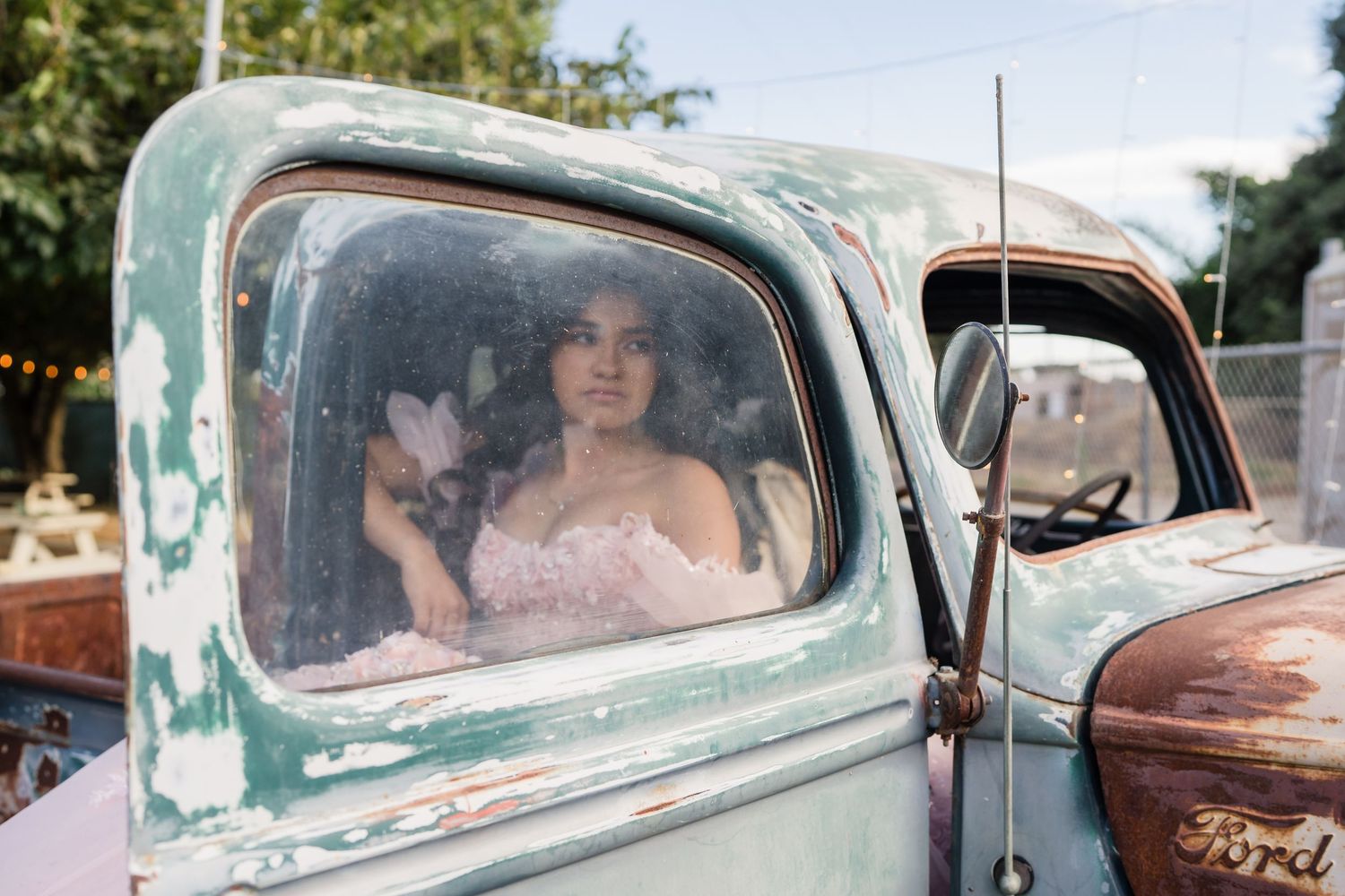 Bride in pink gown sits inside a vintage turquoise Ford truck at an outdoor wedding venue.