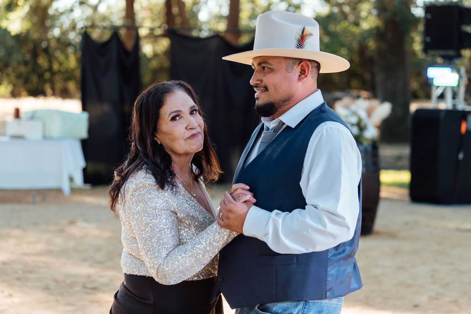 Groom in white cowboy hat and navy vest dances with older woman at outdoor wedding reception.
