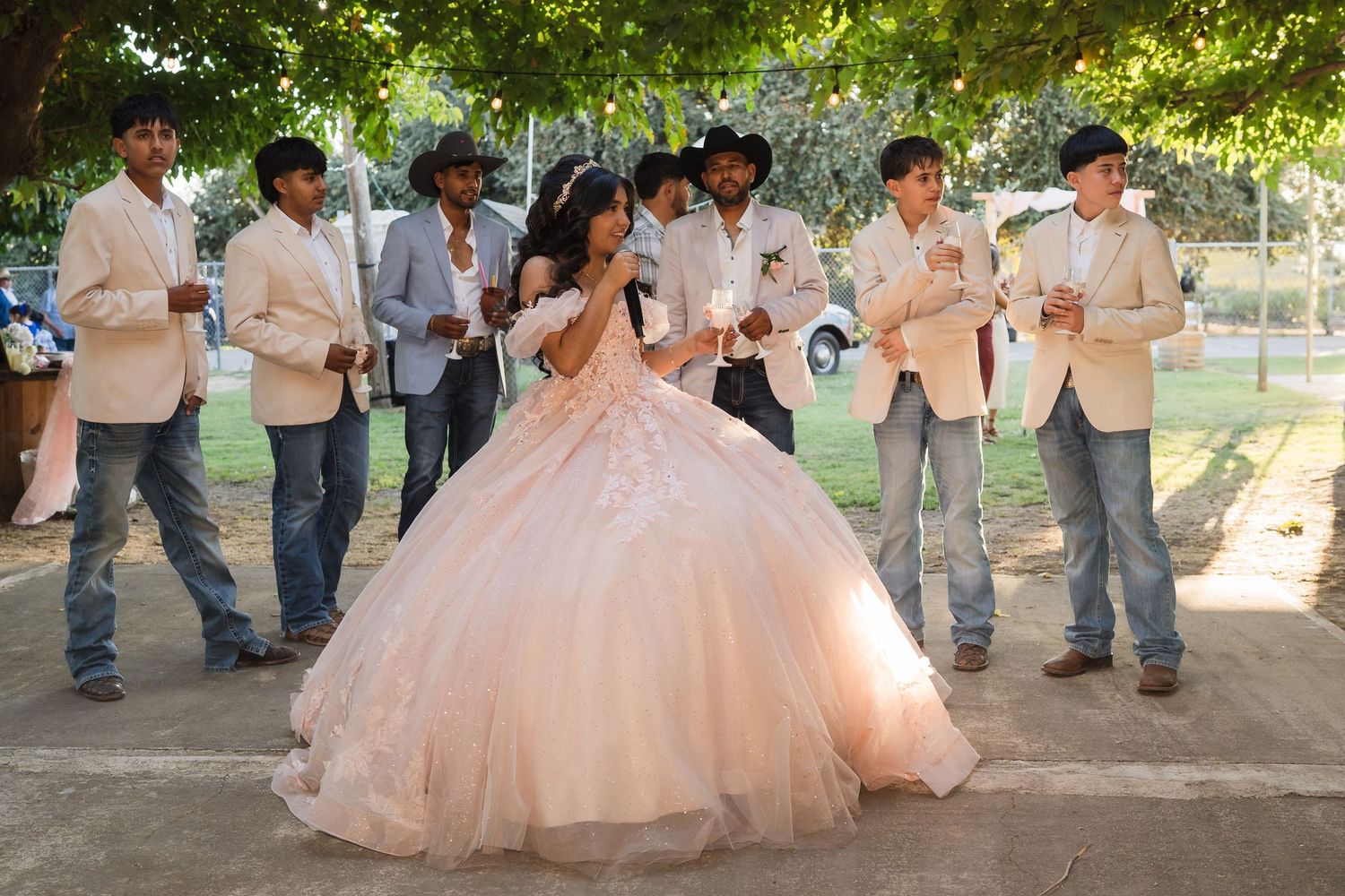 Quinceañera girl in blush ball gown dances with chambelanes in beige suits during outdoor celebration.