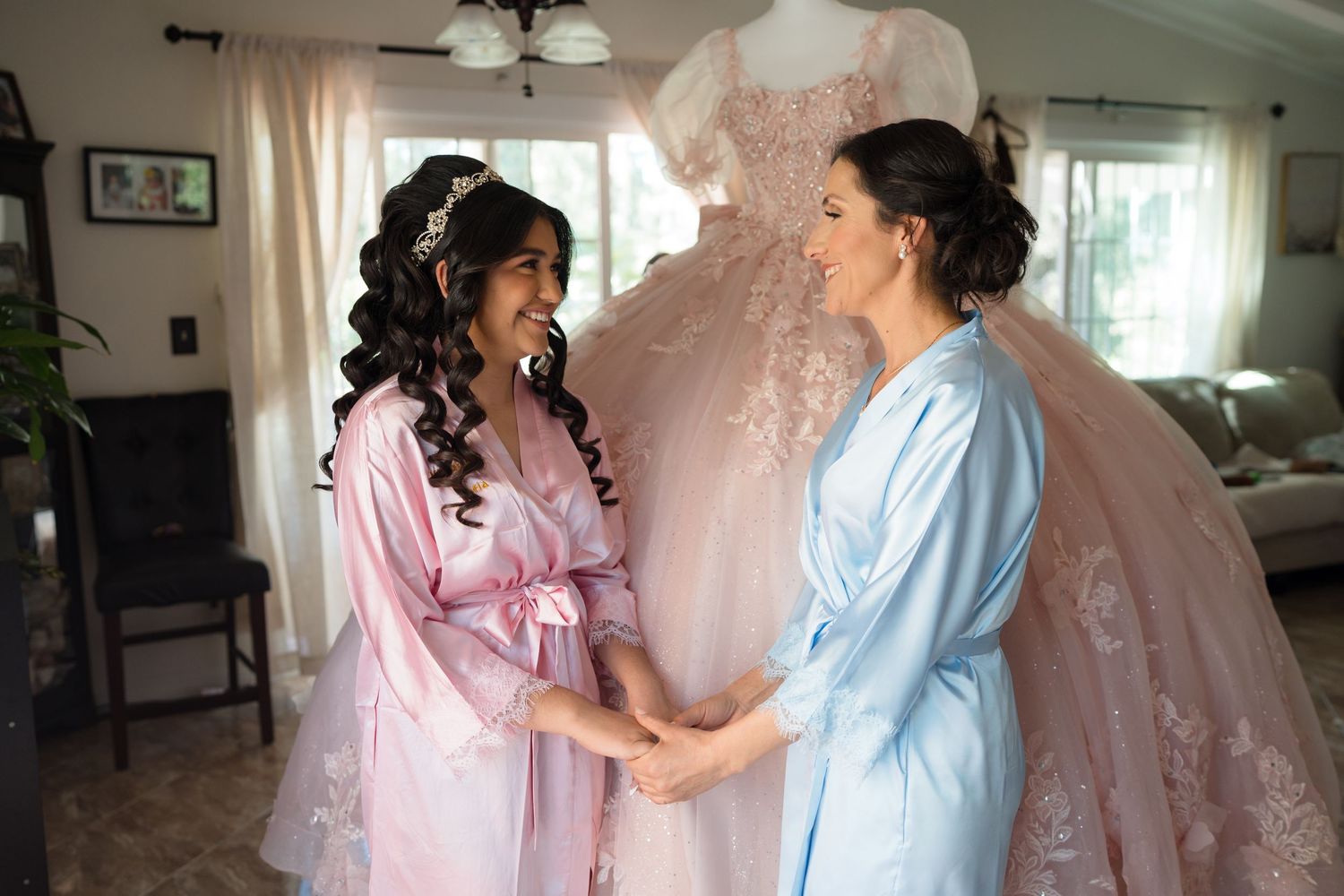 Two women in robes holding hands and smiling before a wedding, with a pink bridal gown displayed in the background.