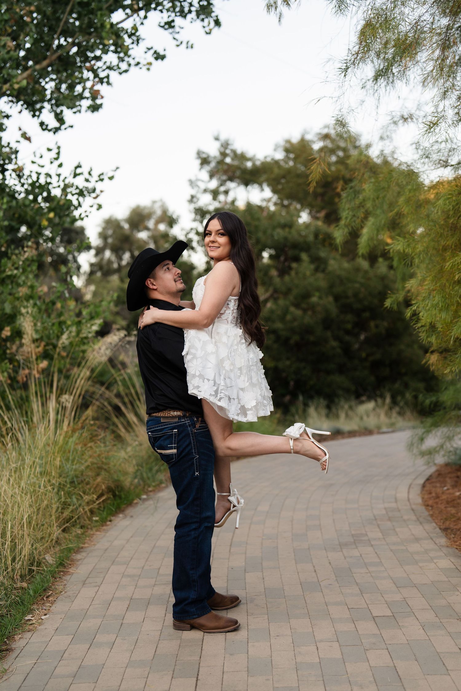 Groom lifting smiling bride in white ruffled dress on a brick pathway surrounded by lush greenery.