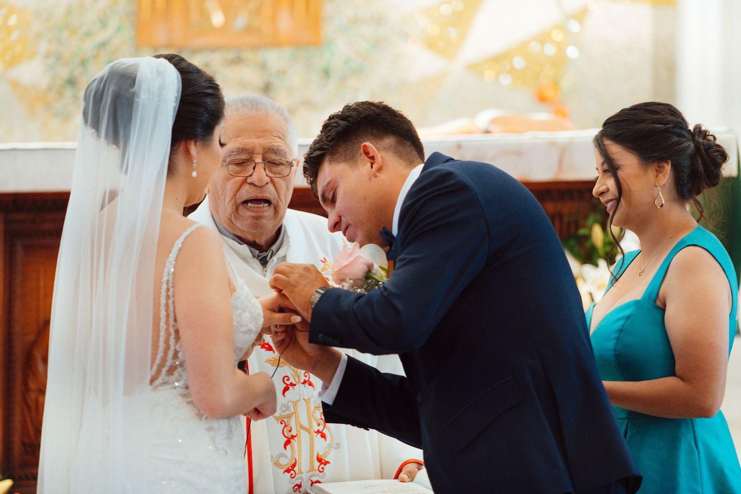 Bride and groom exchange rings with elderly officiant during church wedding ceremony, bridesmaid in teal dress watches.