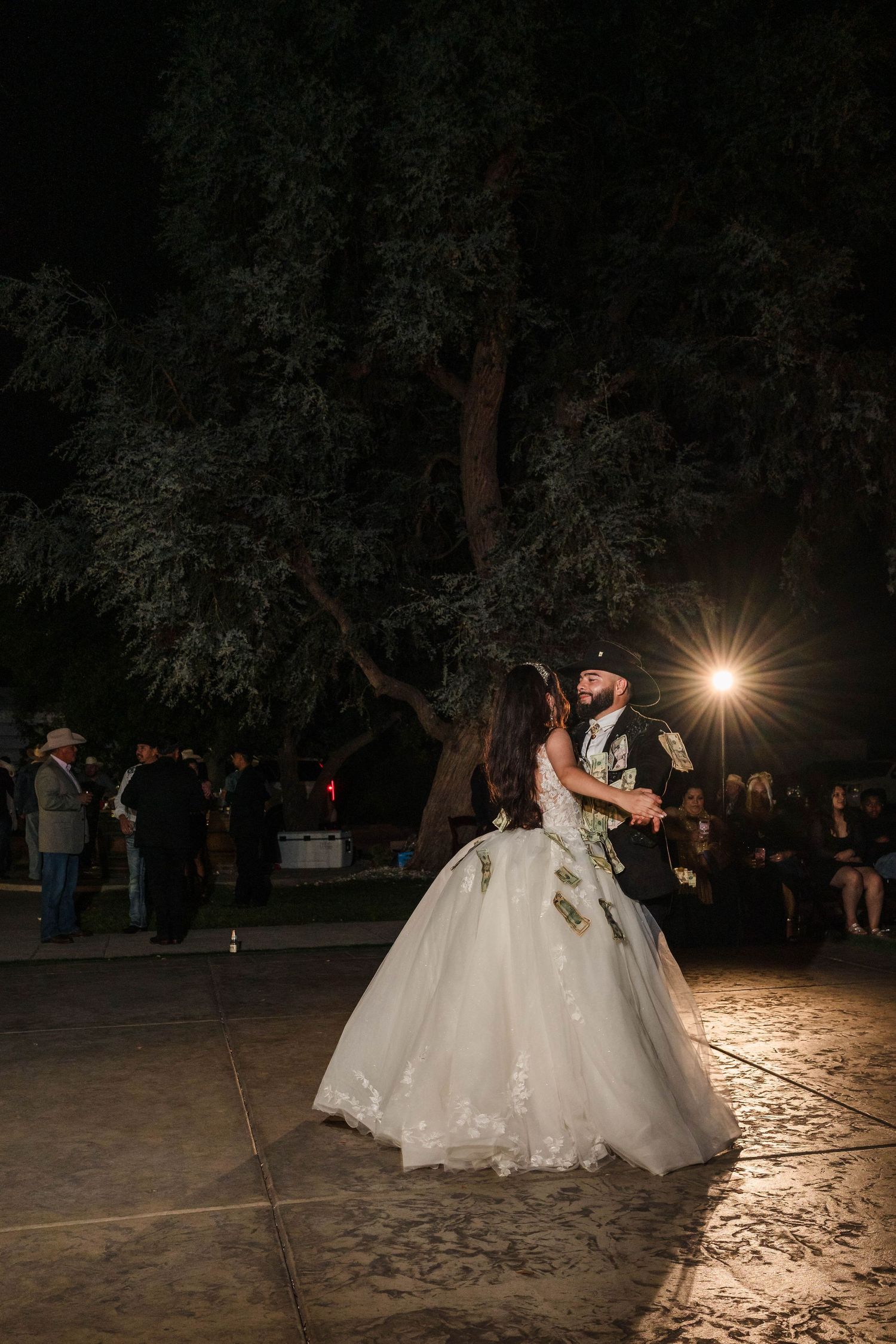 Bride and groom share first dance outdoors at night under trees with warm spotlight at wedding reception.